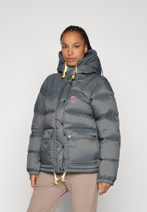 Γκρίζο μπουφάν puffer με ψηλό γιακά, κίτρινες κορδόνες, δύο μπροστινές τσέπες και ορατά κουμπιά. Λαμπερή υφή και σχεδίαση με ραφές.