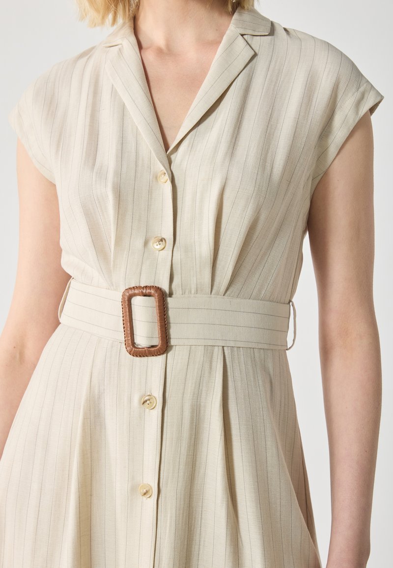 Beżowa sukienka koszulowa w paski z zapięciem na guziki i szerokim pasem w talii z prostokątną brązową klamrą. Gładki materiał, krótkie rękawy.