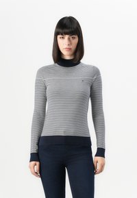 Mujer con cabello oscuro lacio, vistiendo un suéter de rayas azul marino y blanco de manga larga y jeans oscuros, contra un fondo blanco.