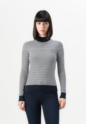 Donna con capelli scuri lisci che indossa un maglione a maniche lunghe a strisce blu navy e bianche e jeans scuri su uno sfondo bianco.