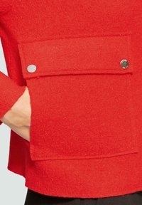 Czerwona wełniana kurtka o teksturowanym wykończeniu, z dużą naszywaną kieszenią zapinaną na napy oraz lekko zaokrąglonym dołem.