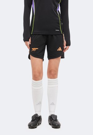 Person trägt schwarze Fußballshorts mit orangefarbenen Arsenal- und Adidas-Logos, weiße knielange Socken mit Adidas-Streifen und schwarze Fußballschuhe.