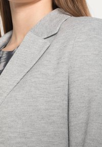 Detailní záběr na osobu v světle šedém texturovaném saku přes vzorovaný šedý top, ukazující krk a oblast ramen.