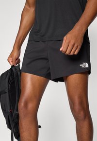 Schwarze Sportshorts mit strukturiertem Muster, ausgestattet mit einem seitlichen Schlitz und dem weißen Logo "The North Face". Ein schwarzer Rucksack wird in der linken Hand gehalten.