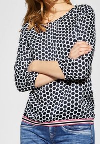 Kvinna med korslagda armar iklädd en blus med svart och vit prickig mönster samt blå jeans, sedd från axlarna till höfterna.