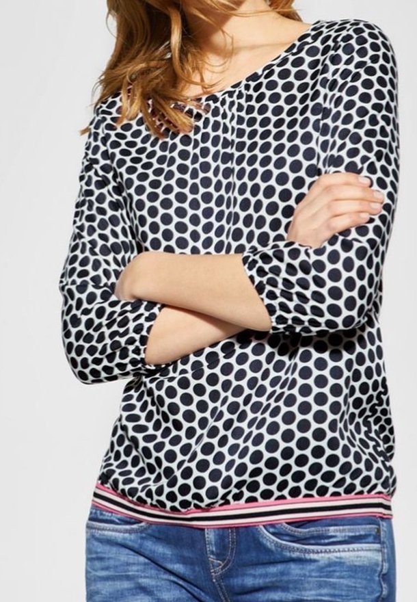 Kvinna med korslagda armar iklädd en blus med svart och vit prickig mönster samt blå jeans, sedd från axlarna till höfterna.