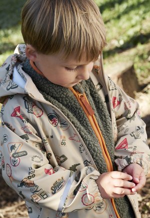 Bambino piccolo con giacca fantasia e gilet in pile guarda verso il basso, tenendo un piccolo oggetto all'aperto sotto la luce naturale del sole.