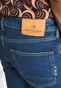 Vaqueros de mezclilla con lavado azul, con un parche de cuero etiquetado "SCOTCH&SODA", costuras amarillas y detalles decorativos bordados en el bolsillo trasero.