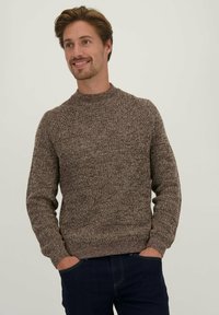 Bruine gebreide trui met een hoge hals, geribbelde manchetten en zoom, met een geperforeerd patroon. Model draagt donkere jeans, handen in de zakken.