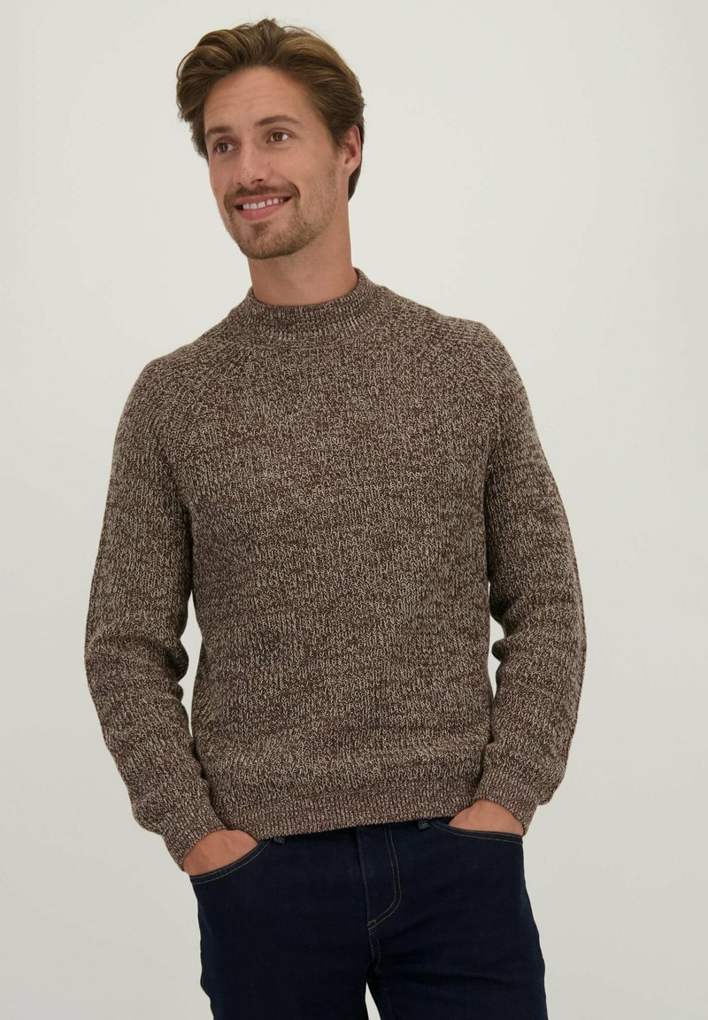 Bruine gebreide trui met een hoge hals, geribbelde manchetten en zoom, met een geperforeerd patroon. Model draagt donkere jeans, handen in de zakken.