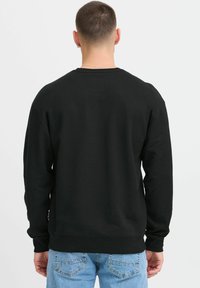 Sudadera negra con cuello redondo, hecha de un tejido suave y texturizado. Presenta mangas largas y un corte relajado, combinada con jeans de color azul claro.