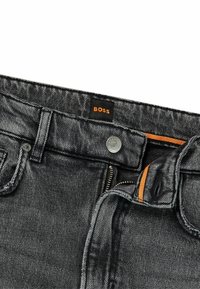 Mustat farkut, joissa on nappikiinnitys, vetoketju ja oranssi yksityiskohta sisävyötäröllä. Ompeleva logo näkyvissä.