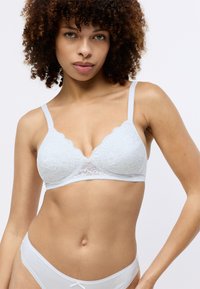 Balta mežģīņu bralette ar viļņotām malām, ar mīkstu tekstūru un trīsstūrveida krūštura kausiem. Apvienots ar atbilstošām baltām zemu vidukļa apakšbiksēm.