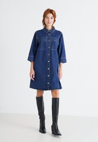 Vestido de camisa de mezclilla en azul oscuro con cuello, botones en la parte delantera y dos bolsillos en el pecho. Lleva unas botas negras hasta la rodilla, con punta afilada.