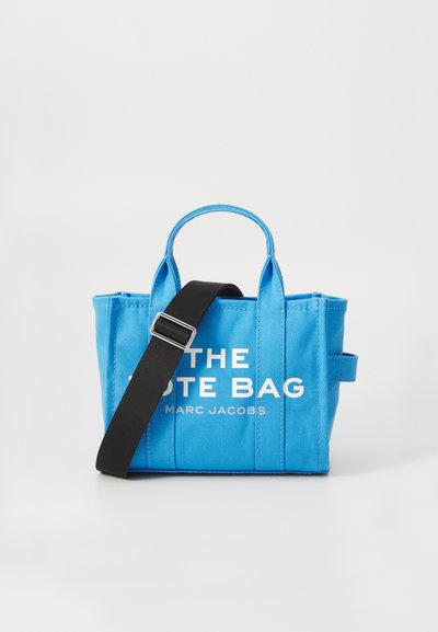 Sac fourre-tout rectangulaire bleu avec une bandoulière réglable noire, des poignées courtes et le texte blanc "THE TOTE BAG MARC JACOBS" sur le devant.