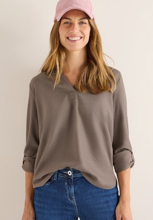 Chemise marron à manches longues avec un col en V et des poignets retroussés, associée à un jean en denim bleu. Portée avec une casquette en velours côtelé rose, texture lisse.