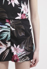 Černý květinový overal s velkými růžovými a bílými květy a zelenými listy, vyrobený z hladkého materiálu. Zahrnuje přiléhavý top a šortky.