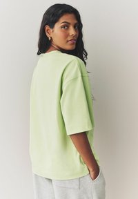 T-shirt oversize vert clair à manches courtes, col rond et coupe décontractée, porté avec un pantalon de survêtement gris, sur un fond neutre.