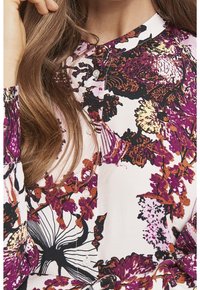 Blusa de estampado floral en blanco, con vibrantes diseños en púrpura, negro y naranja. Tiene un cuello redondo y detalles de botones.
