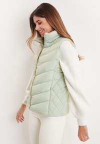 Hellgrüne, gepolsterte Weste mit einem gesteppten Design, hohem Kragen und einem Reißverschluss vorne. Getragen über einem weißen, strukturierten Pullover.