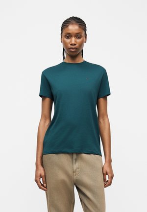 Frau mit geflochtenem Haar, die ein dunkelgrünes kurzärmeliges T-Shirt und beige Hosen trägt, steht vor einem schlichten weißen Hintergrund.