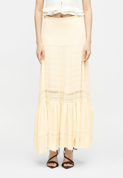 Γυναίκα που φοράει κρεμ maxi φούστα με στρώσεις, ψηλή μέση και λεπτομέρειες από δαντέλα, μαζί με μαύρα ψηλοτάκουνά σανδάλια με λουράκια.