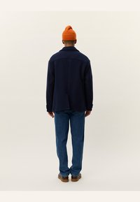 Chaqueta de mezcla de lana en color marino con un corte relajado, que presenta una costura cosida en la parte trasera y una tapeta delantera, combinada con unos jeans de mezclilla azul y un gorro naranja.