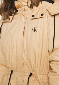 Beige parka with a drawstring waist, black accents, snap buttons, and a logo. Material appears smooth with a slightly glossy finish.