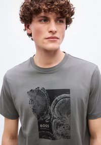 Νέος με σγουρά μαλλιά, που φοράει γκρι μπλουζάκι με εκτυπώσεις εγκάρσιων τομών δακτυλίων δέντρου σε μαύρο και τη λέξη "BOSS" στη μπροστινή πλευρά.