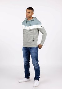 Sudadera gris claro con franjas horizontales en teal y blanco, cordones, bolsillo frontal; combinada con jeans azules y zapatillas blancas.