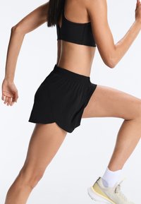 Fekete sportmelltartót és rövidnadrágot viselő nő fut, fehér zoknit és bézs tornacipőt hord, oldalról látható fehér háttér előtt.