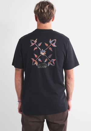 T-shirt en coton noir arborant un motif floral et géométrique coloré au dos, avec le nom de la marque "OXBOW" en police courbée.