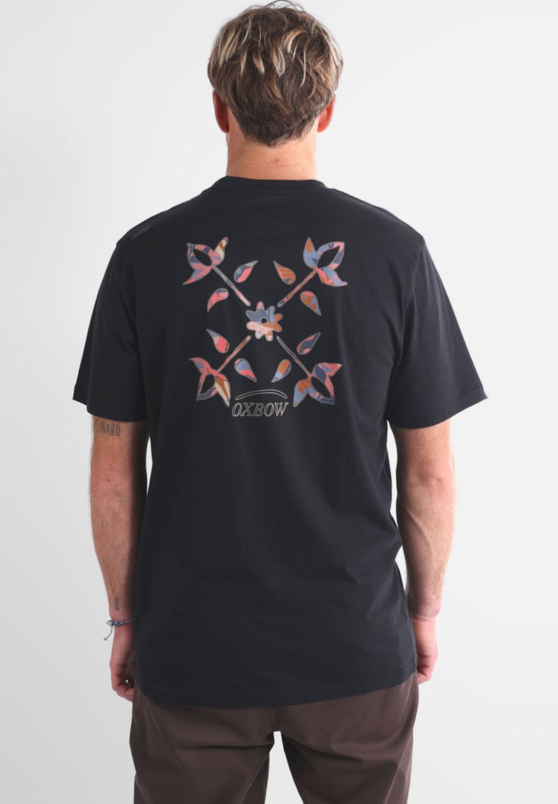 T-shirt en coton noir arborant un motif floral et géométrique coloré au dos, avec le nom de la marque "OXBOW" en police courbée.