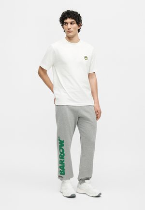 Homme debout portant un t-shirt blanc avec un petit logo smiley jaune et un pantalon de survêtement gris avec le texte vert "BARROW" sur la jambe gauche.