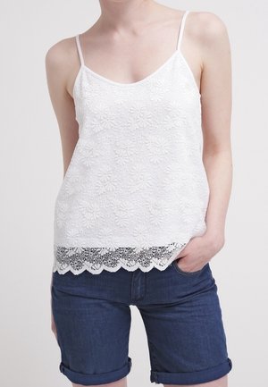 Žena v bílém květovaném krajkovém tílku s tenkými ramínky a tmavě modrých džínových šortkách s ohrnutými nohavicemi, ruka v kapse.