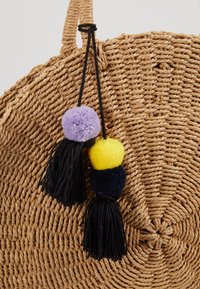 Sac rond en paille tressée avec anses tressées et pompons décoratifs noirs avec pompons violet, jaune et bleu foncé suspendus à une anse.