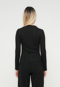 O femeie cu părul lung și blond poartă un top negru cu dungi, ajustat, cu mânecă lungă și pantaloni negri, stând pe un fundal simplu deschis.