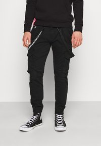 Svart cargo-bukser med sidelommer, justerbare stropper og grafisk tekstdetalj. Brukt med svarte høyhælte sneakers med hvite snøringer.