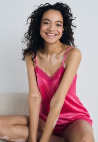 Roze satijnen topje met kantafwerking, voorzien van spaghettibandjes en een gladde textuur. Model poseert zittend met ontspannen armen.