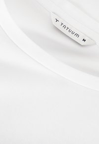 Bílý bavlněný tričko s kulatým výstřihem, hladkou texturou a štítkem s nápisem "TNTUUM M" na vnitřním límci.