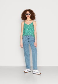Top de tanque floral verde com alças finas, combinado com jeans azul claro e sapatilhas brancas. Modelo de pé contra um fundo liso.