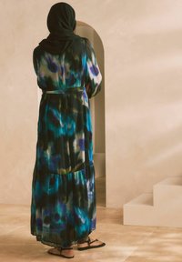 Robe longue et fluide avec un motif aquarelle bleu, vert et noir. Présente un design à volants et une ceinture assortie. Des sandales à lanières sont portées.
