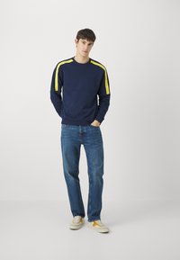 Felpa blu marino con strisce gialle sui lati, collo a giro e polsini a costine, abbinata a jeans blu e scarpe con accenti gialli.