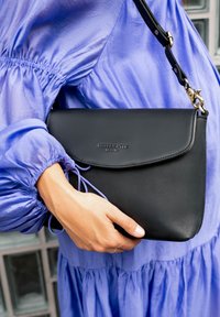 Person in blue dress holding a black leather crossbody bag with a flap and metal clasp, embossed "HOLZRCHTER BERLIN" on front.