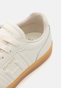 Vita lädersneakers med guld "HACKETT"-logotyp, beige snören och en ljusbrun gummisula. Har diskreta designinslag.