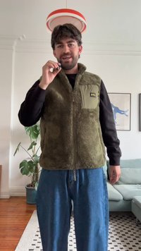 Grüne Fellweste mit Reißverschluss und einem aufgesetzten Taschen. Getragen über einem schwarzen Hemd und kombiniert mit lockeren Jeans.