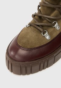 Parte superior de ante oliva con acentos de cuero burdeos, puntera redonda, suela de goma marrón resistente con ranuras horizontales y ojales de metal para los cordones.