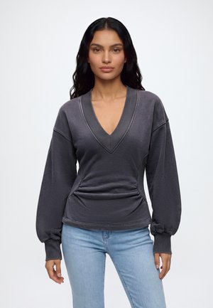 Femme aux longs cheveux foncés portant un pull gris foncé à col en V avec des manches ballon et un jean bleu clair, debout devant un fond uni.