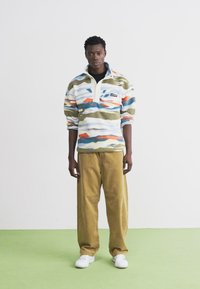 Pullover in felpa con motivo a onde multicolore, base bianca, colletto con half-zip; abbinato a pantaloni di velluto a coste color marrone chiaro e sneakers bianche.