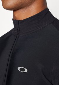 Black athletic jacket featuring a high collar, textured accents, and a slim fit. It has a silver logo on the front for branding.
